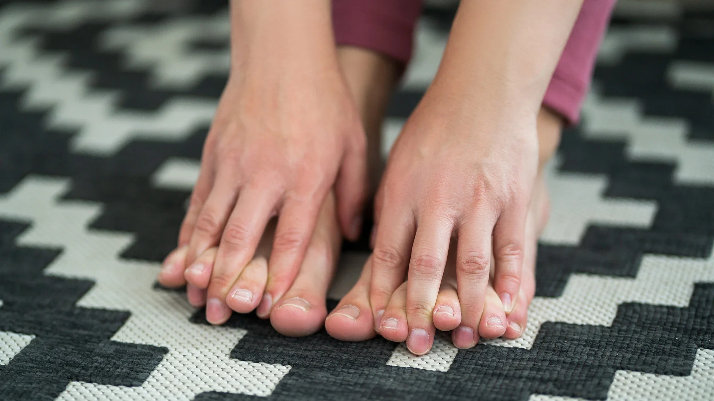 Close-up of a person reaching town to rub their feet. Their fingers are interlaced in between their toes. There is a black and white patterned rug beneath their feet.
Staras/iStock via Getty Images