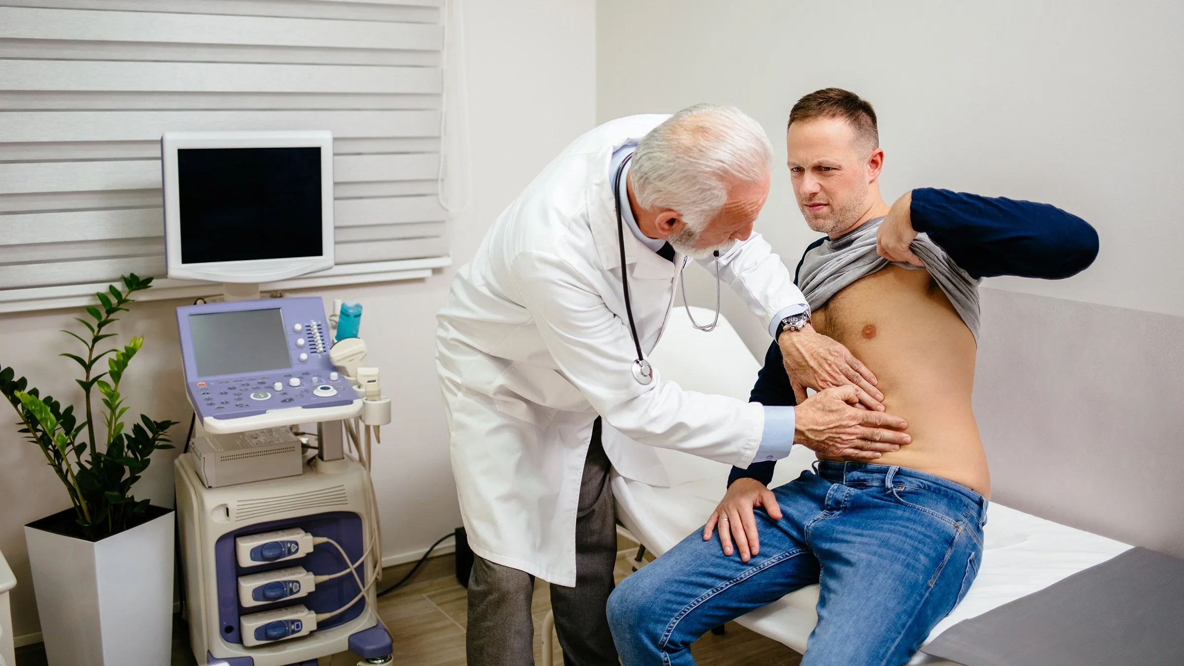 Doctor examining a patient's abdomen. He is pressing down on the upper intestine area to check for pain. The patient is making a slight squinting face in discomfort.
