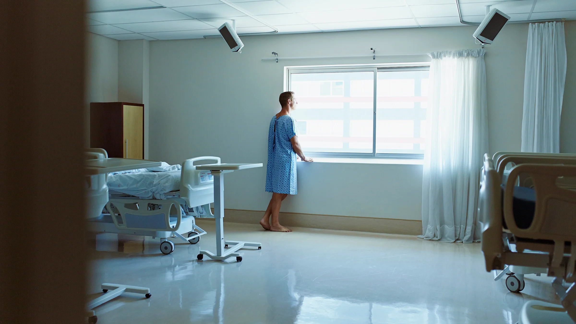 Man in hospital gown staring out the hospital room window. The room is empty of any other people.