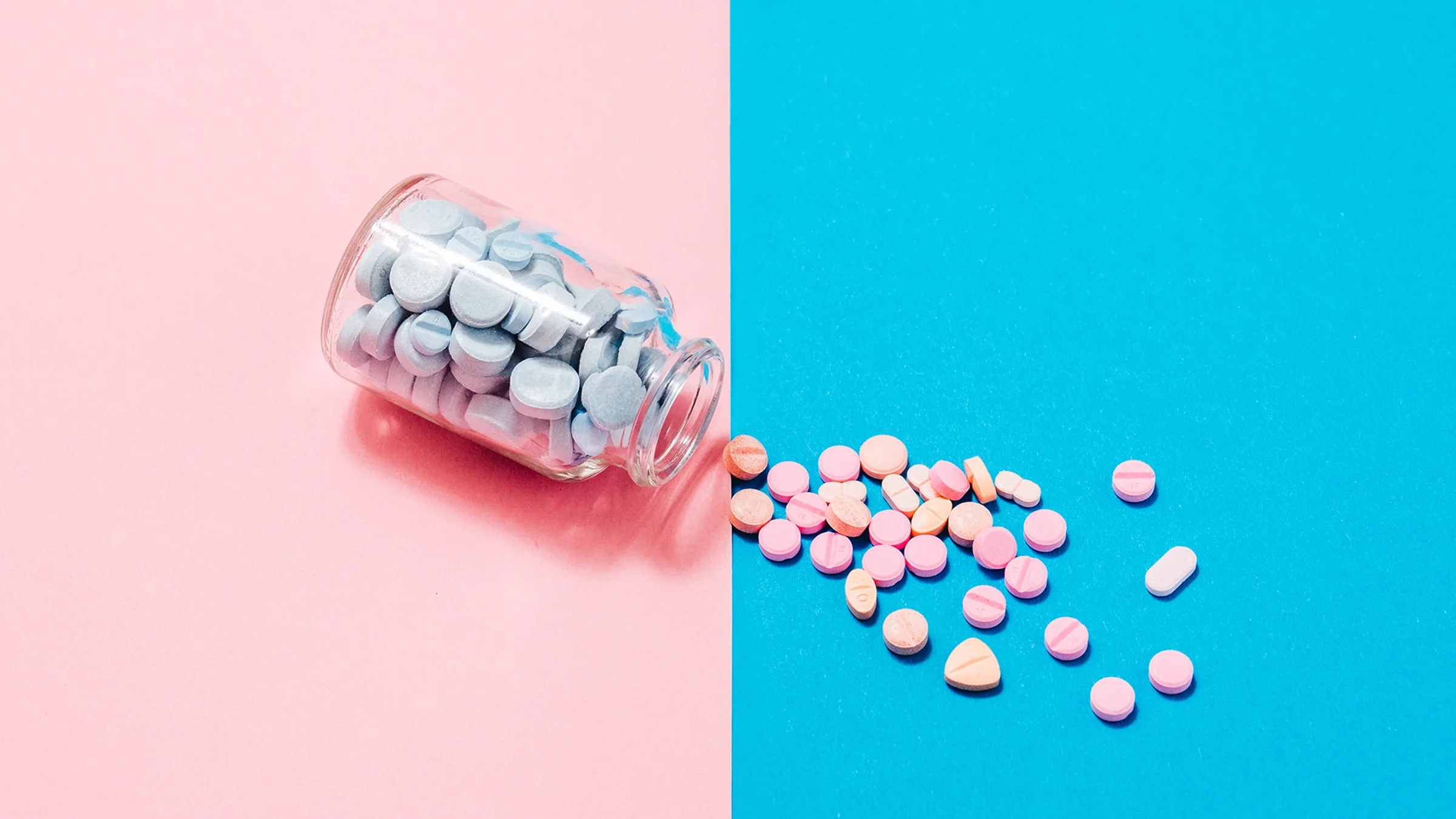 The image depicts a glass jar of blue pills lying on its side against a pink background, and pink pills spilling out onto a blue background.