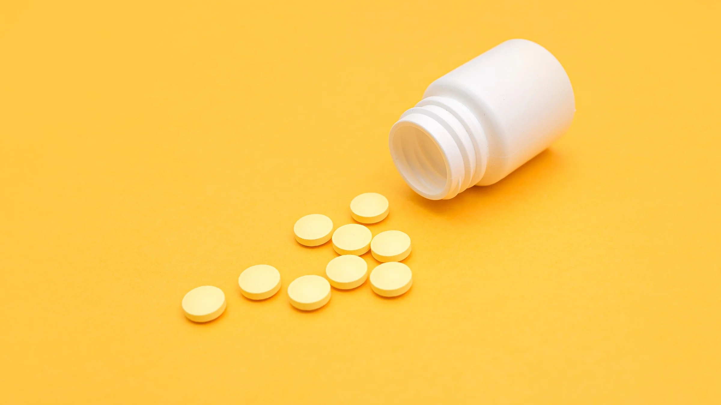 White pill bottle on it's side showing the spilled out pills on a bright yellow background. There are 9 pills that are yellow and round.