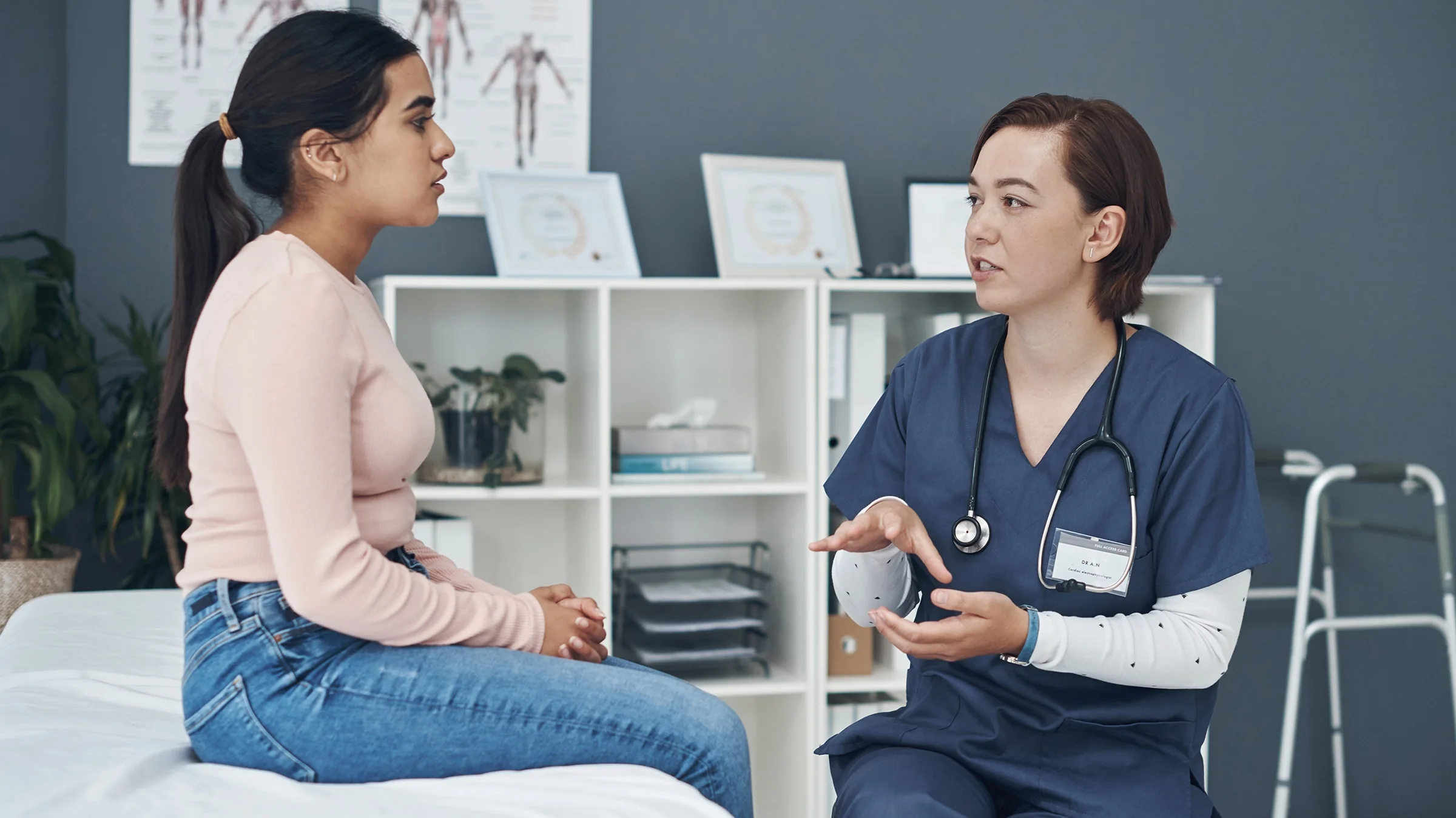 A woman is talking with a nurse in an exam room. The woman is sitting up on the exam table and the nurse is lower on a stool.