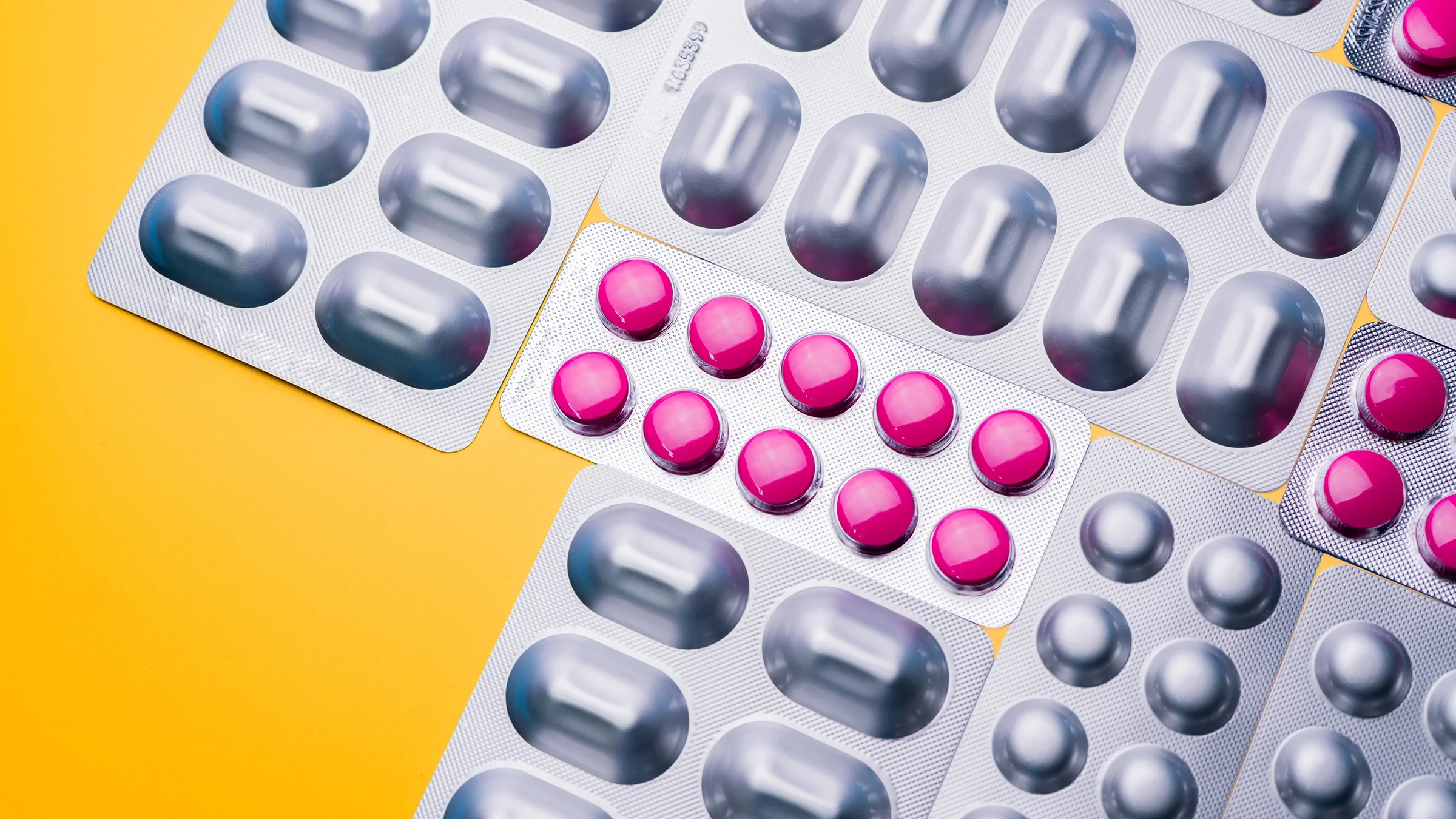 Silver blister packs of pills, some with pink pills visible all on a yellow background.