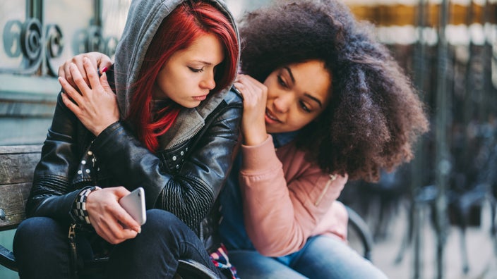 A girl hugs her friend in distress on the front stoop of a home.