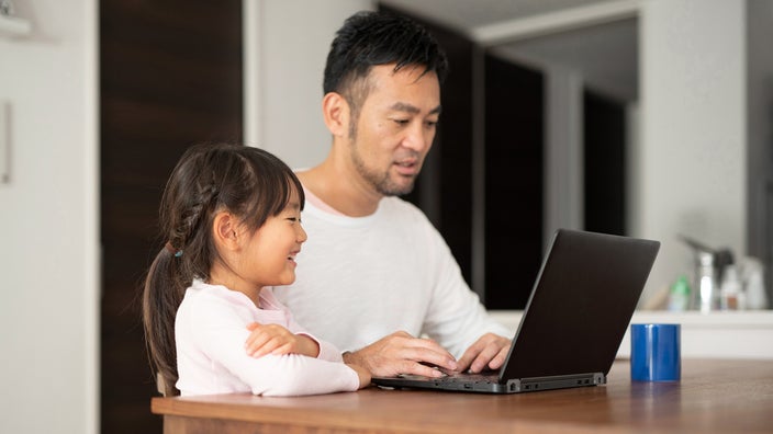 A father sitting at his laptop working while his young daughter stands beside him looking over his shoulder and smiling.