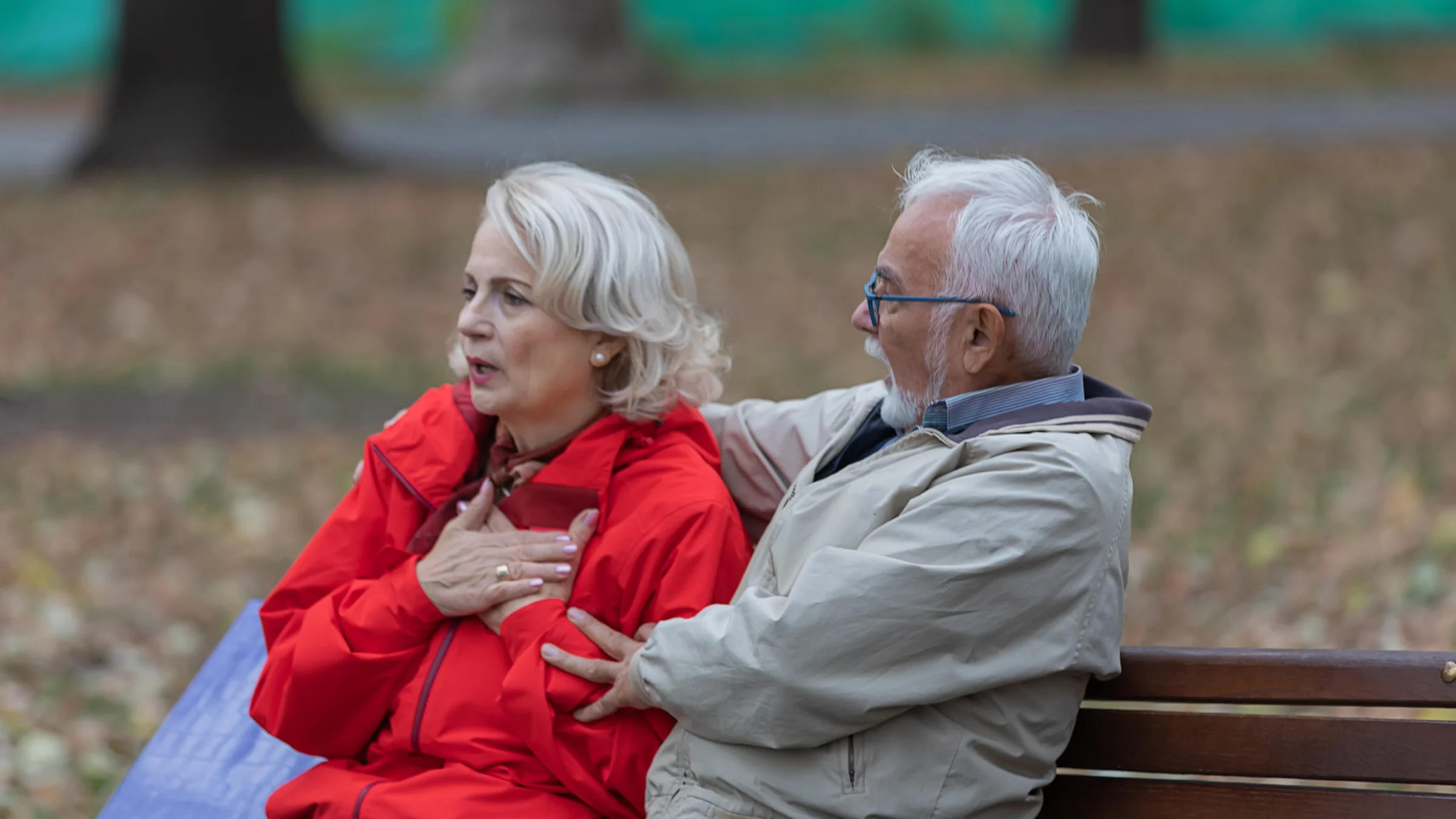An older couple is sitting on a bench in the park. The woman has her hands over her chest and her partner is looking at her.