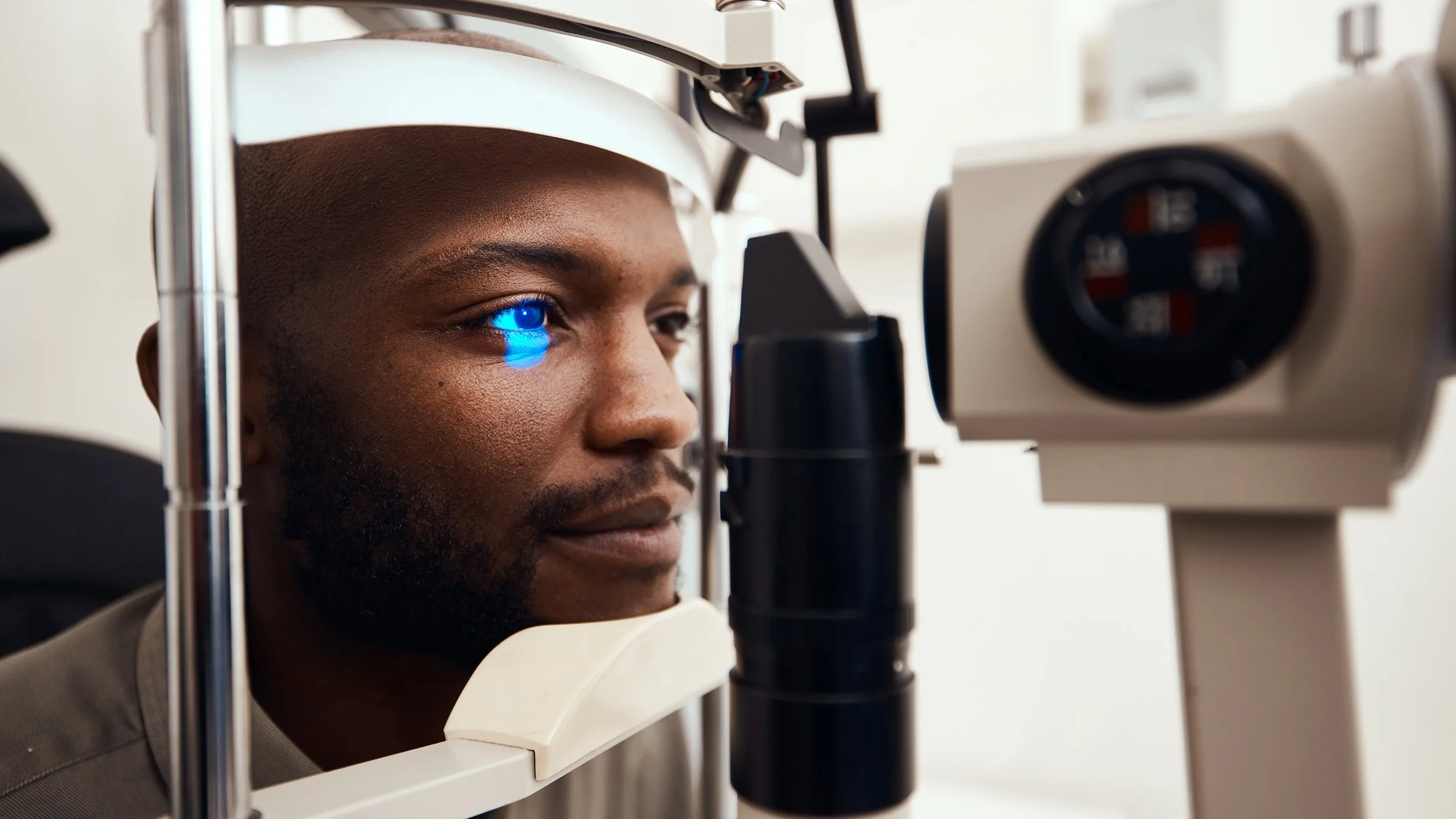 Close-up of a man at the eye doctor. The doctor is using a machine to look into his eye. The man's chin and forehead are resting on the machine and there is a bright blue light shining into one eye.