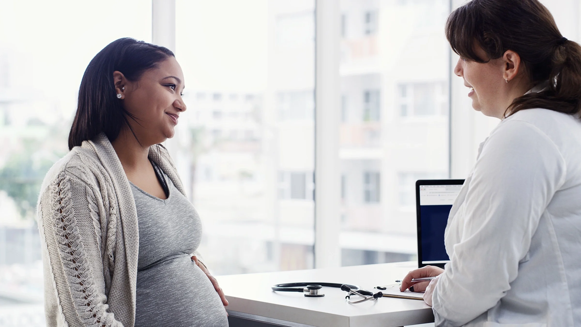 Pregnant woman talking with her doctor in the office. She is softly smiling and listening attentively while touching her belly.