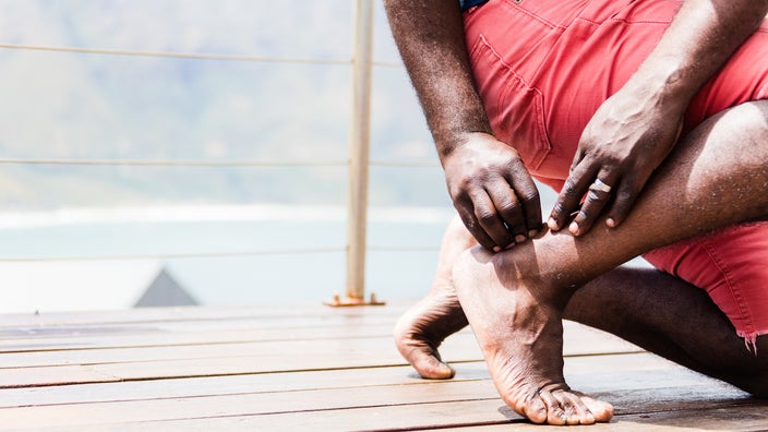 Cropped shot of a man bending down to rub the back of his ankle. He is barefoot and walking on a pier near the ocean.