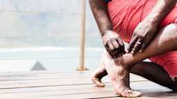 Cropped shot of a man bending down to rub the back of his ankle. He is barefoot and walking on a pier near the ocean.
Jan-Otto/iStock via Getty Images