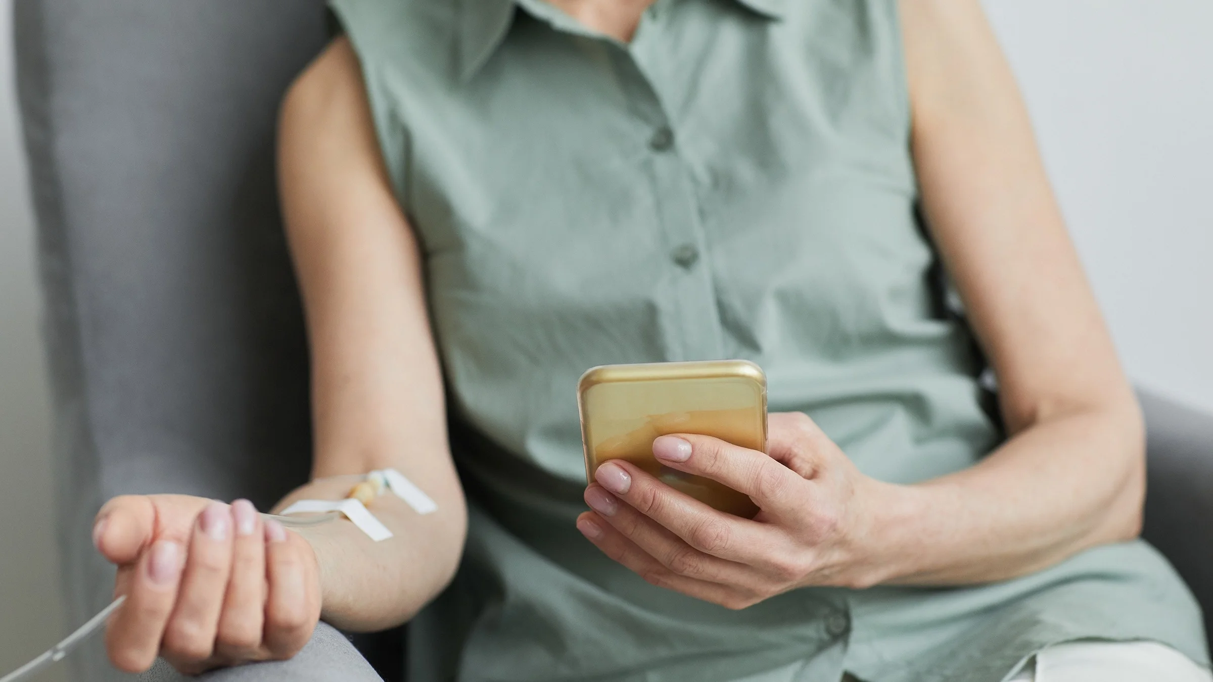 Cropped close-up shot of a woman with an IV in her forearm. She is sitting in a chair and using her mobile phone.