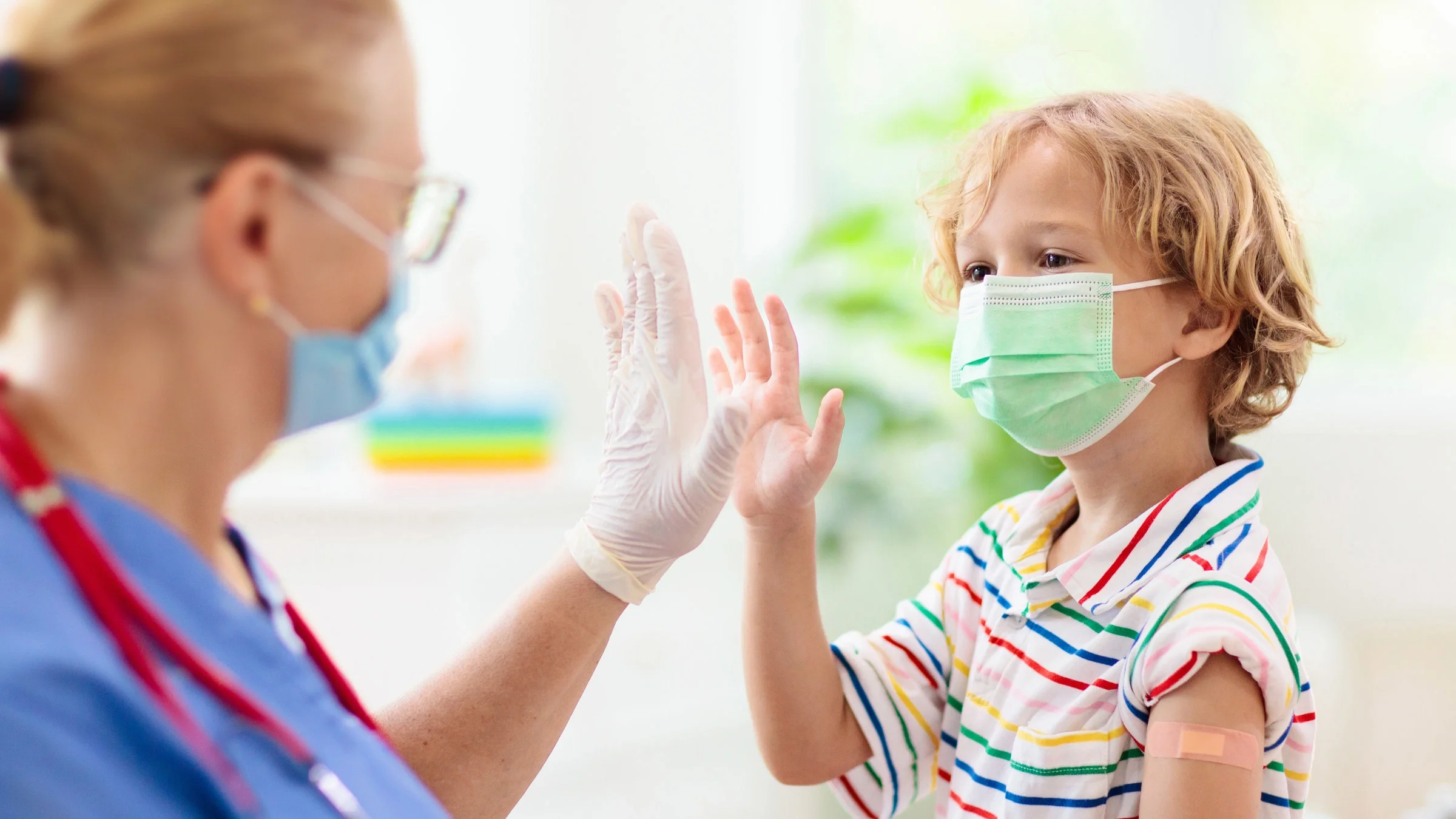 A masked child with a vaccine bandaid on their arm high-fiving their doctor.