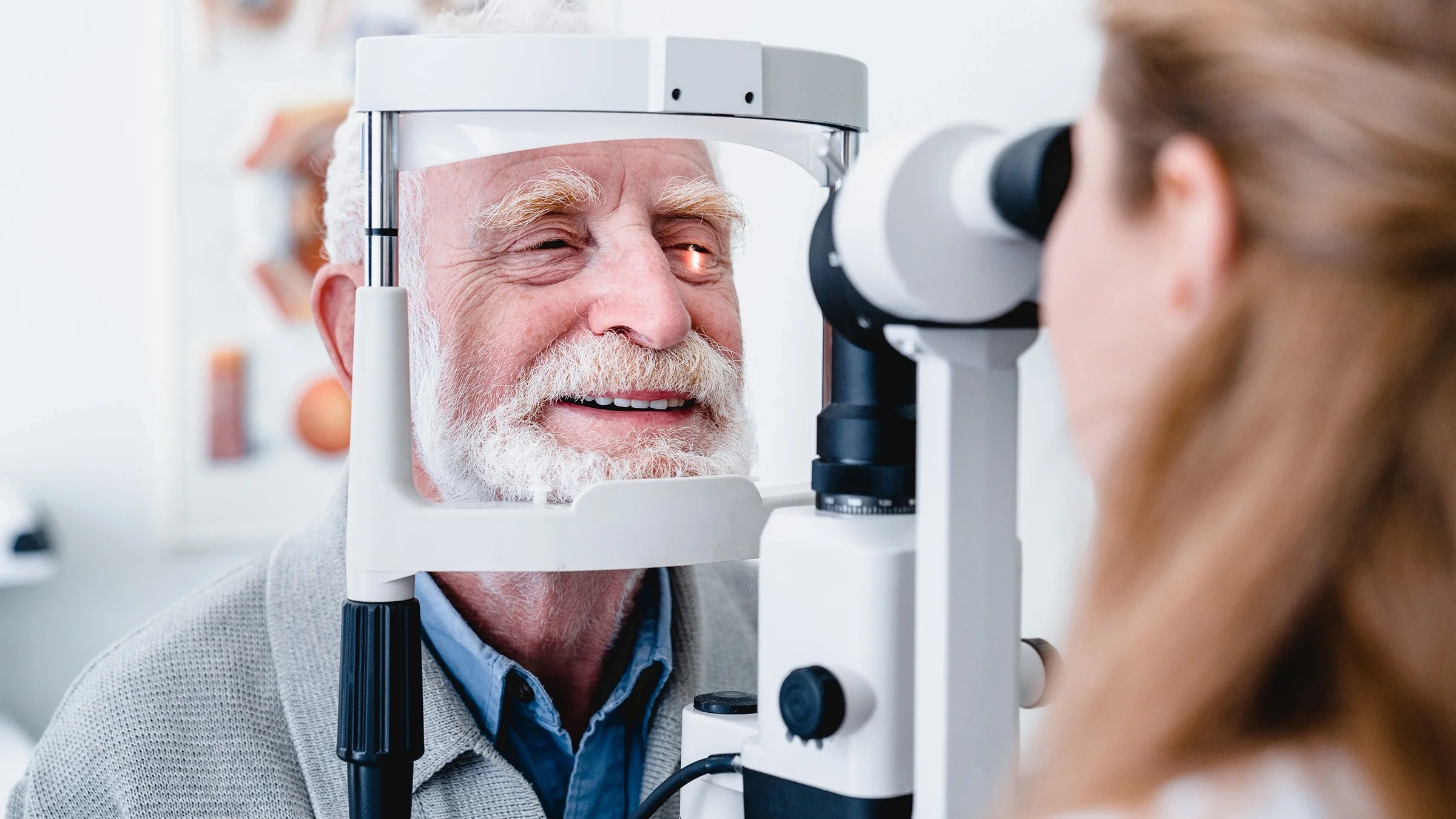 Hombre mayor con el pelo blanco revisándose los ojos en el optometrista. Está apoyando la barbilla y la frente en el equipo de examen y sonriendo.