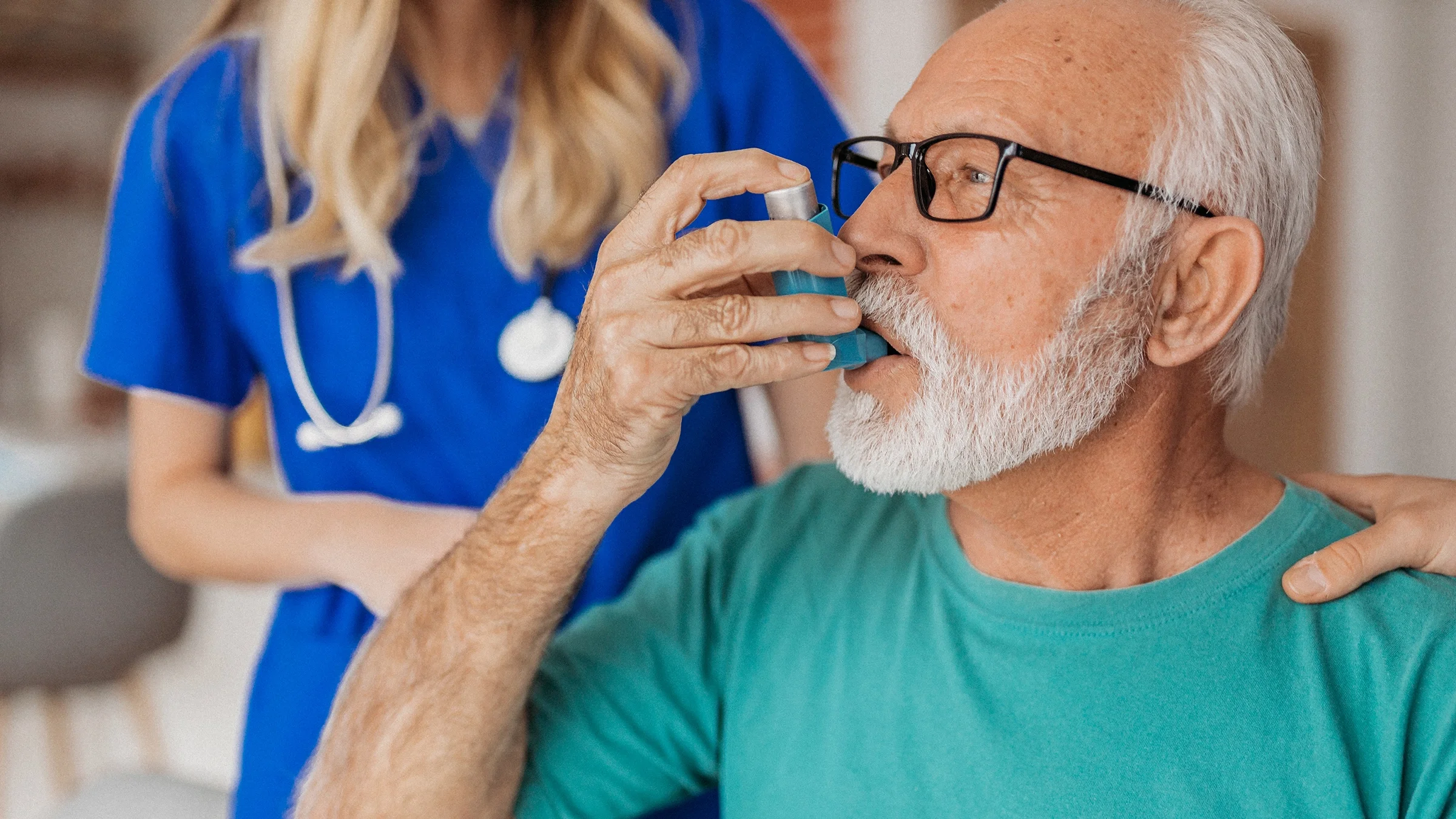 Close-up of a senior man using an inhaler. His nurse in in the background helping him.