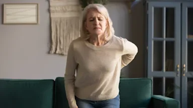 Older woman standing next to the couch clutching her back in pain.
fizkes/iStock via Getty Images