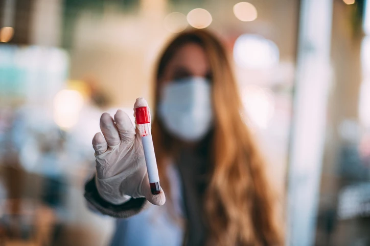Lab technician holding a blood sample with a red cap, the sample is in focus and the person is in the background out of focus.