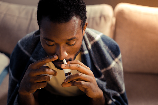 Close up of a young Black man using nose spray in one nostril while sitting on a couch with a blanket wrapped around his shoulders.