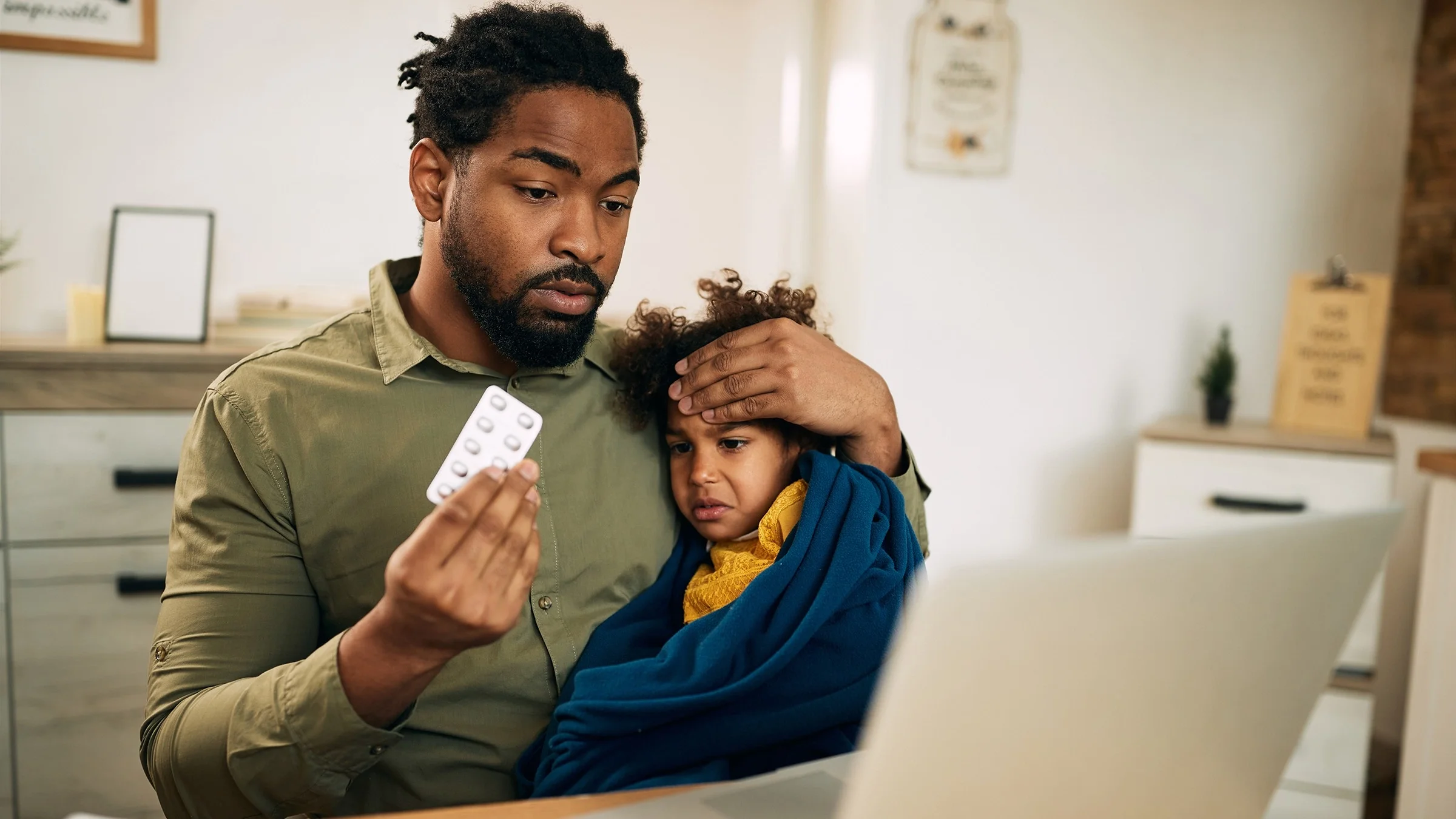 Father holding his sick child in his lap as he is talking to a doctor on a telehealth visit. He is holding a blister pack of antibiotic pills and has a concerned look on his face.