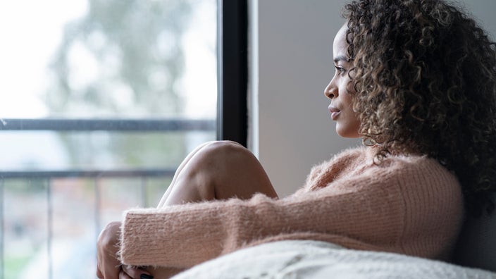 Sad woman with curly hair looking out window. She has her arms wrapped around one of her knees.