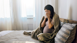 A pregnant woman is blowing her nose while in bed.
skynesher/E+ via Getty Image
