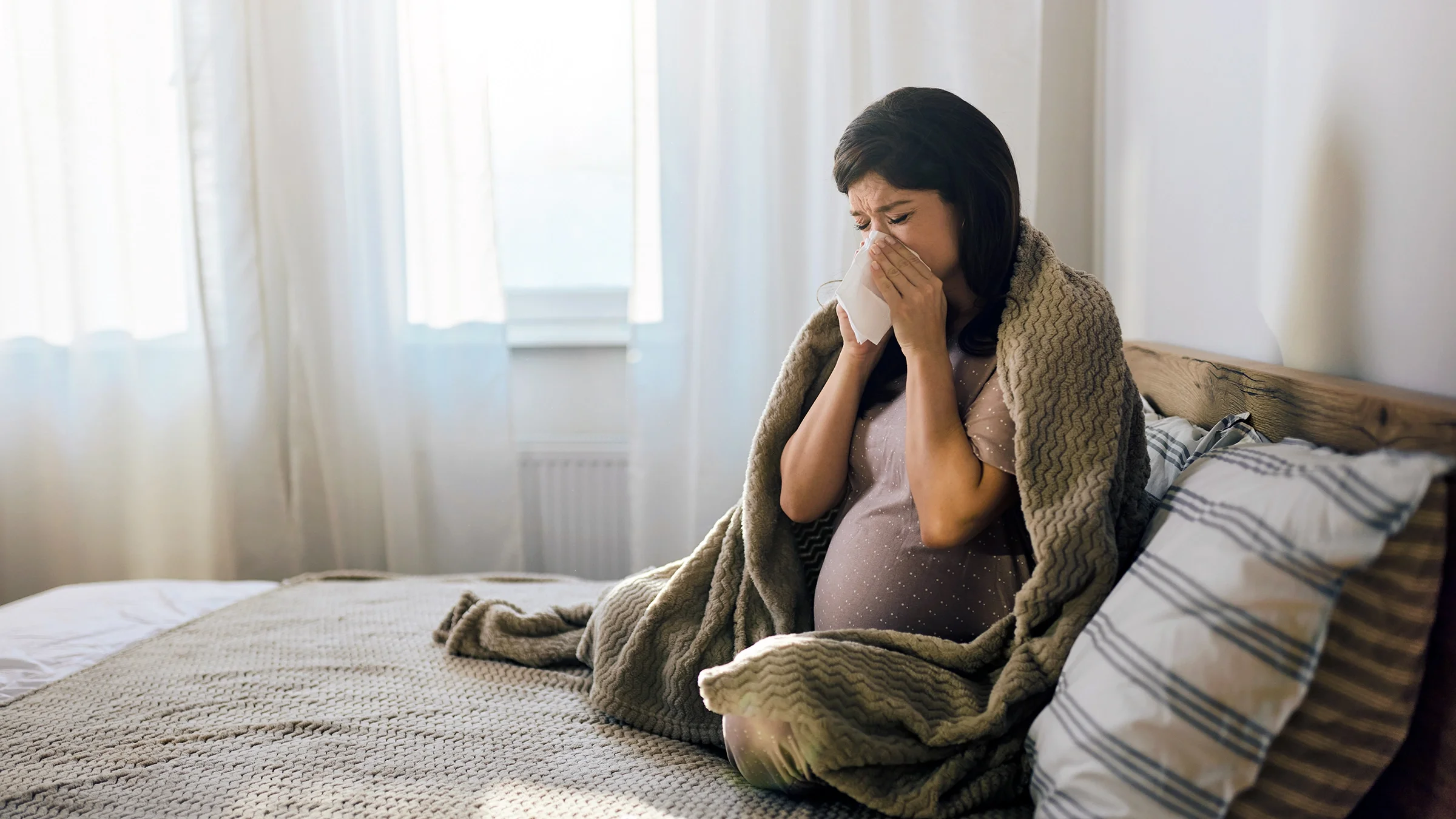 A pregnant woman is blowing her nose while in bed.