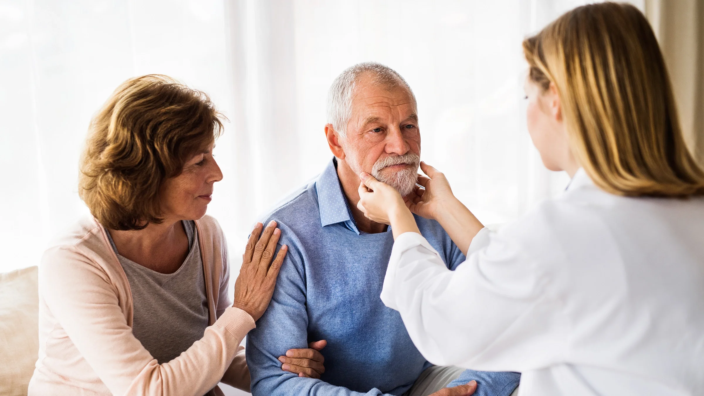 A provider is examining the jaw and chin of an older man. His spouse is with him as well.