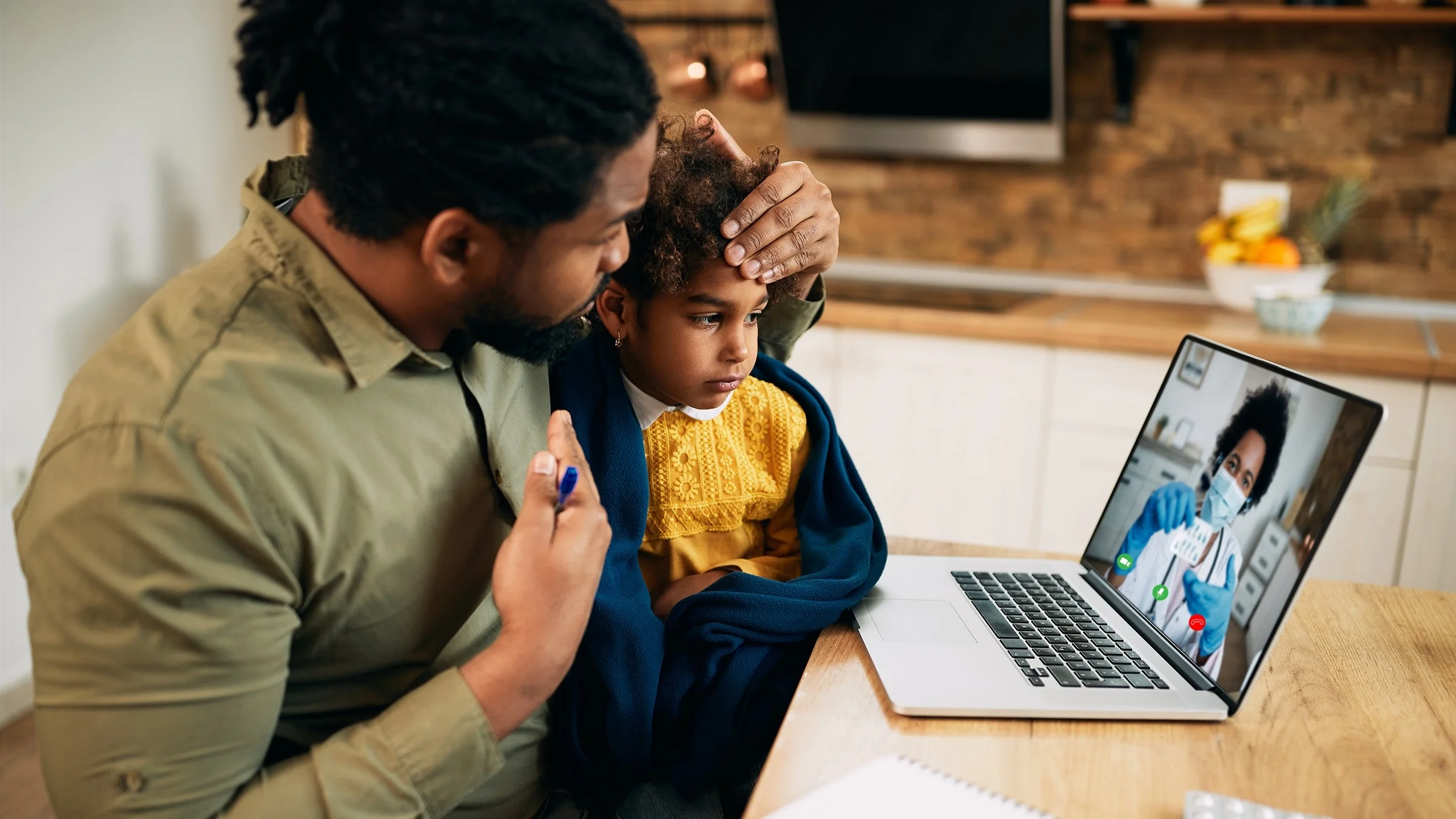 Father holding his daughter on his lap and feeling her forhead for her temperature. They are sitting at the dinning room table on their laptop on a telehealth visit.