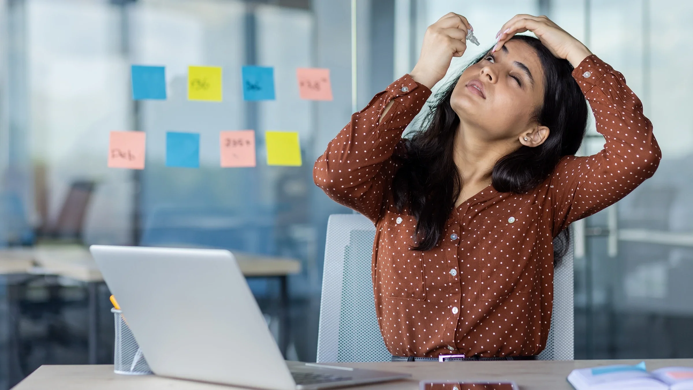 A woman uses eye drops at her desk.
