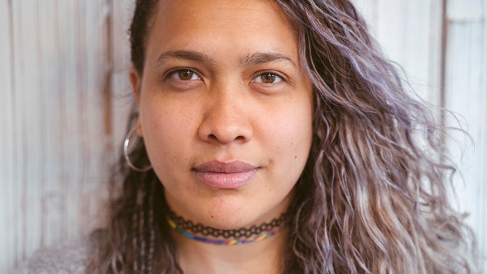 Close-up portrait of a young woman with curly purple gray-ish hair. She is wearing a black choker necklace and has a slight smile on her face.