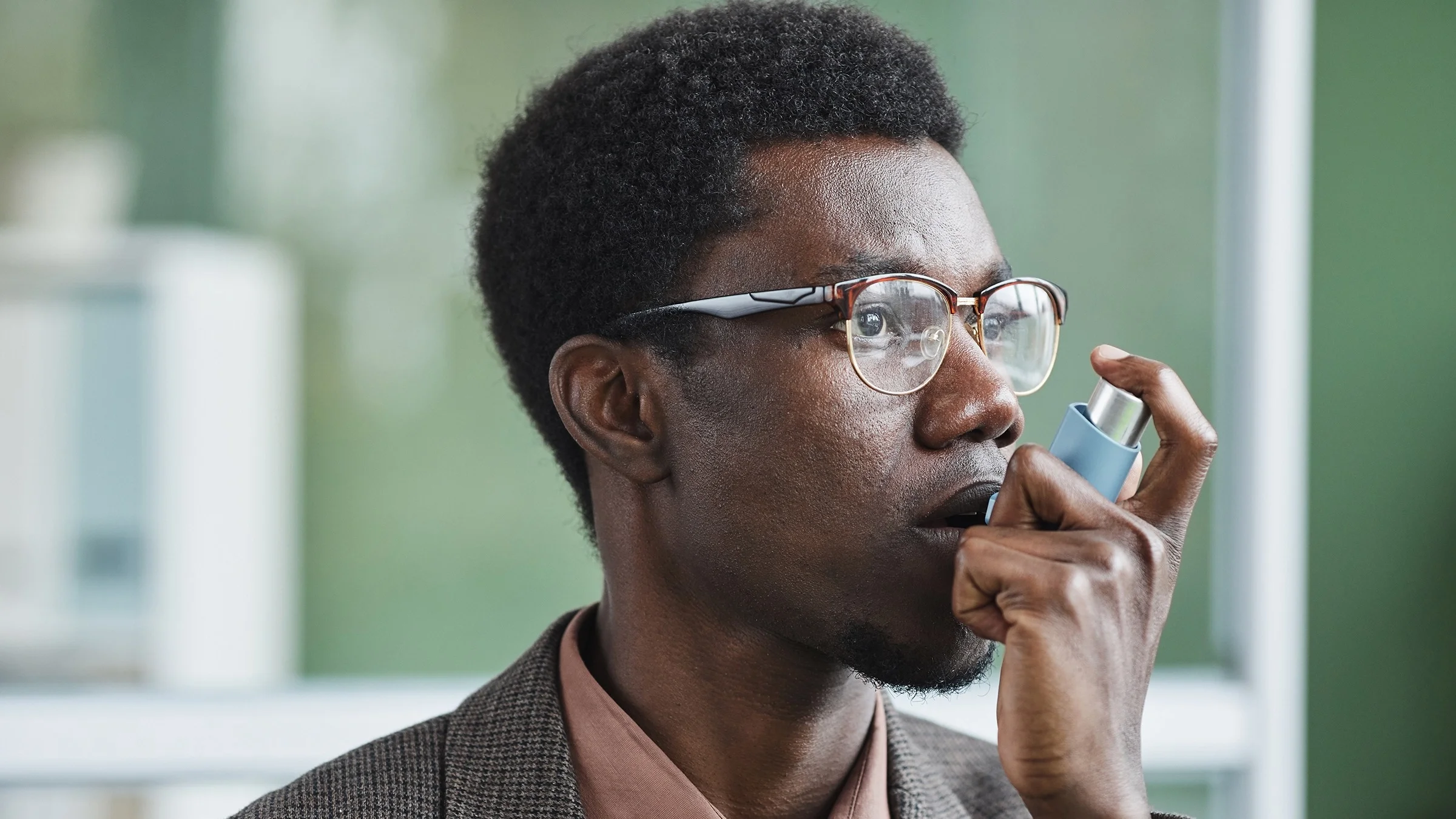 Portrait of a Black man using an inhaler. He is wearing tortoise shell half rimmed glasses and business clothes.