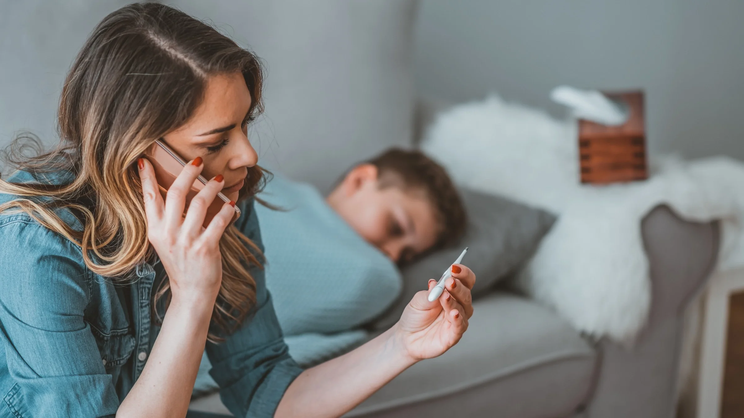 Mother on the phone with the pediatrician as she is looking at a thermometer. You can see her child in the background sleeping on the couch.