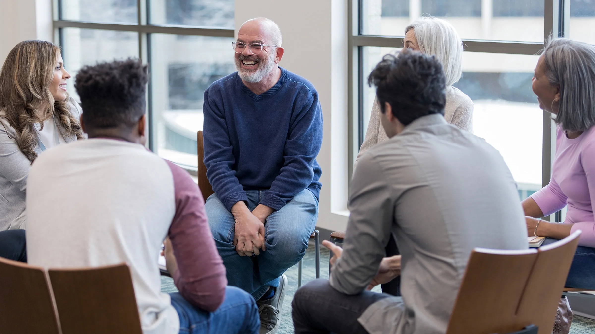 Support group circled up in a large room. The focus is on an older bald man smiling.