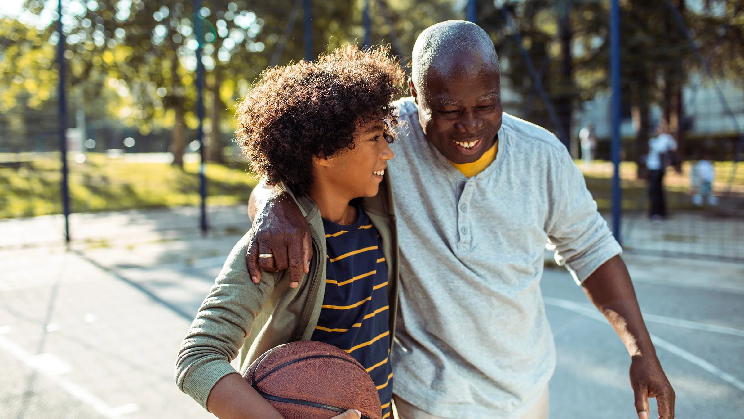 Grandpa and grandson playing basketball outdoors. The grandpa has his arm around his grandson and they are both smiling.