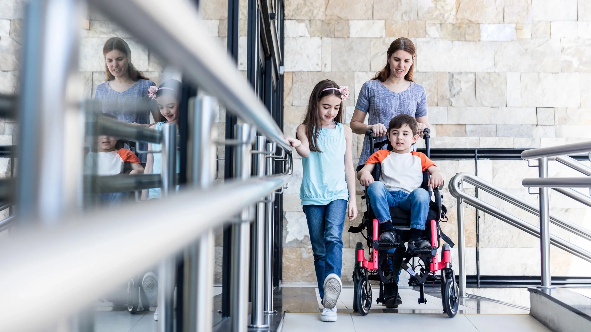Mother walking with her children leaving a medical building. She is pushing her son in a wheelchair and her daughter is walking next to them holding on to the hand rail.