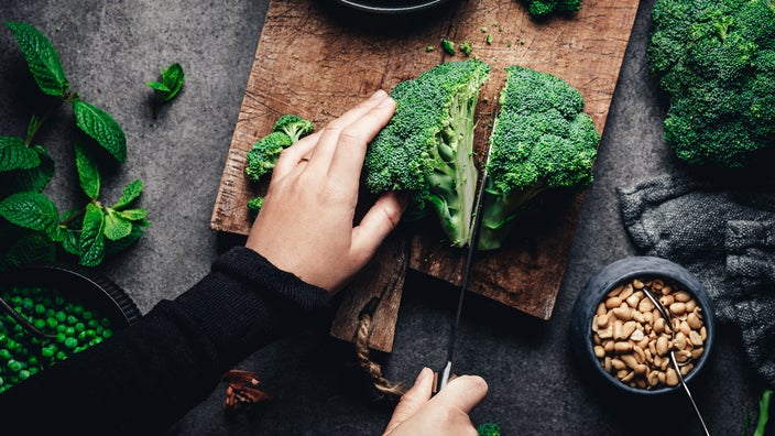 Top down view of a person cutting a broccoli in half on a dark brown cutting board surrounded by other green vegetables and herbs with a textured black table.