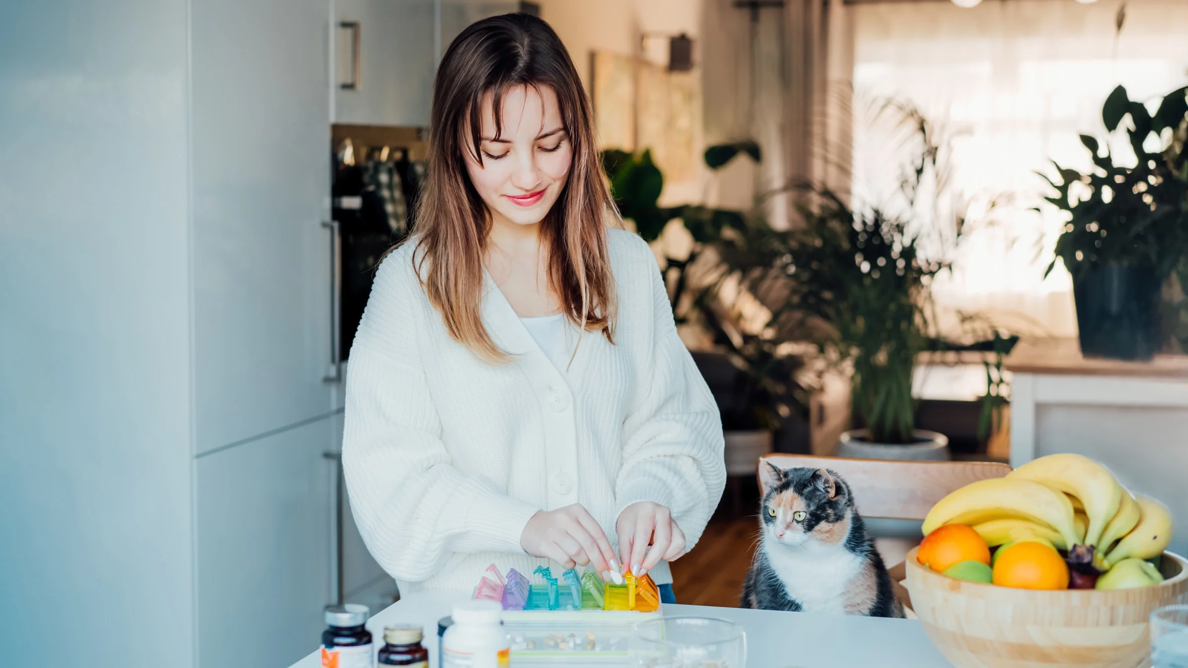 A young adult organizing their pill container at their kitchen counter, next to their cat.