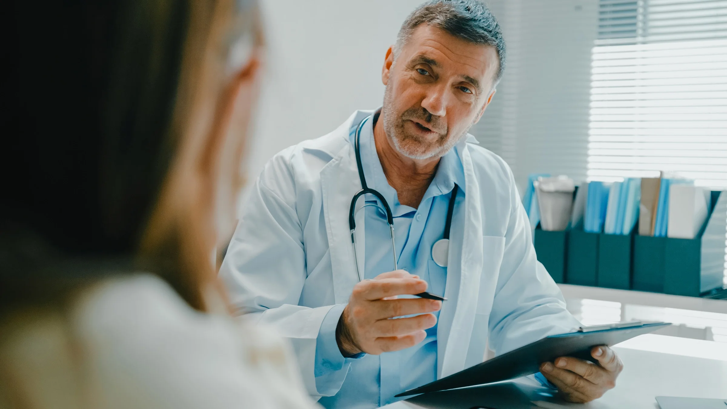 A medical professional, holding a clipboard, speaks with a patient.