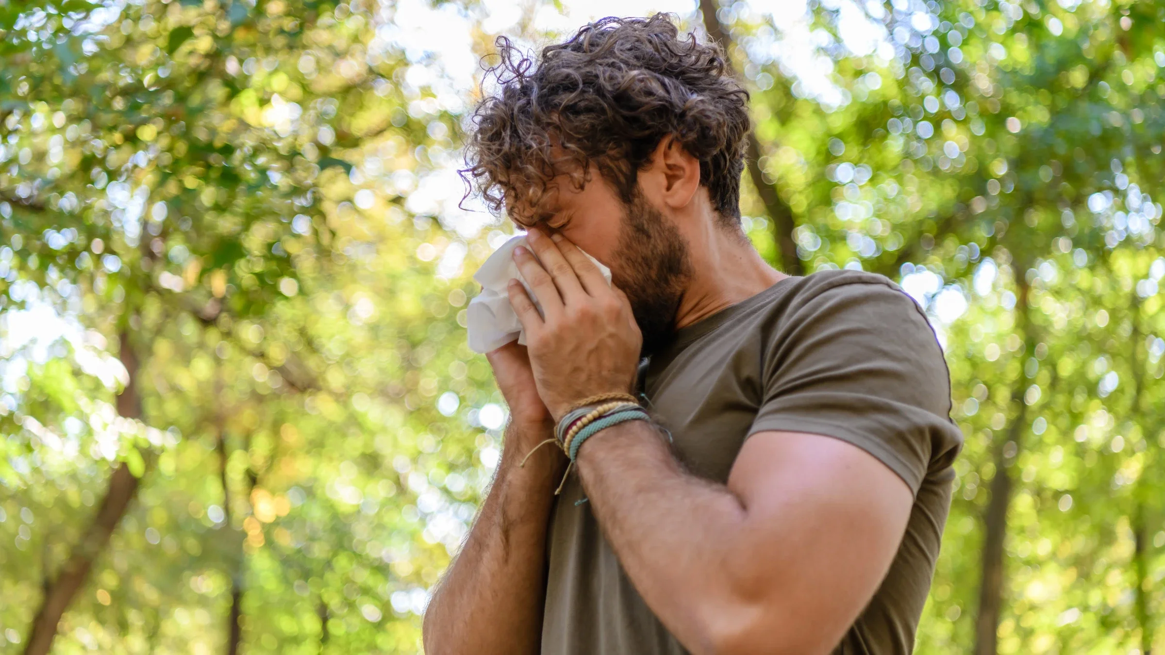 Photo of a man standing in nature. He is sneezing into a tissue. 