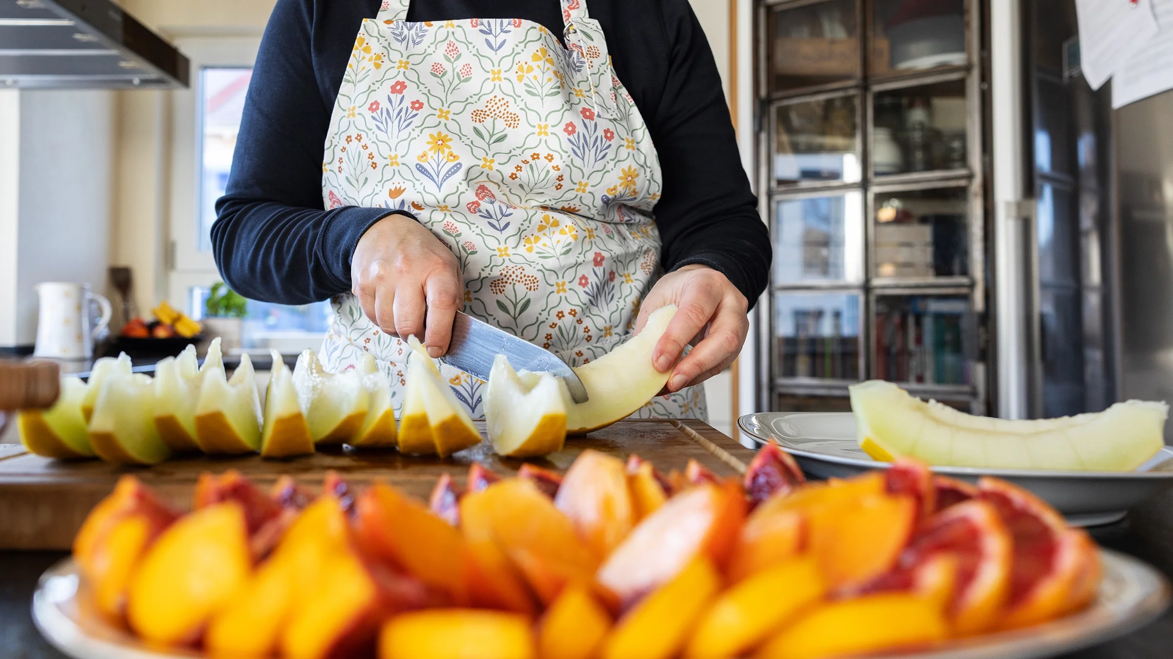 A close-up image shows a woman in the kitchen wearing an apron while slicing melon.