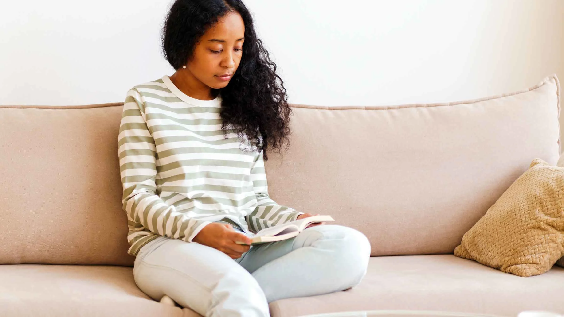 A woman is shown reading a book on a couch at home.