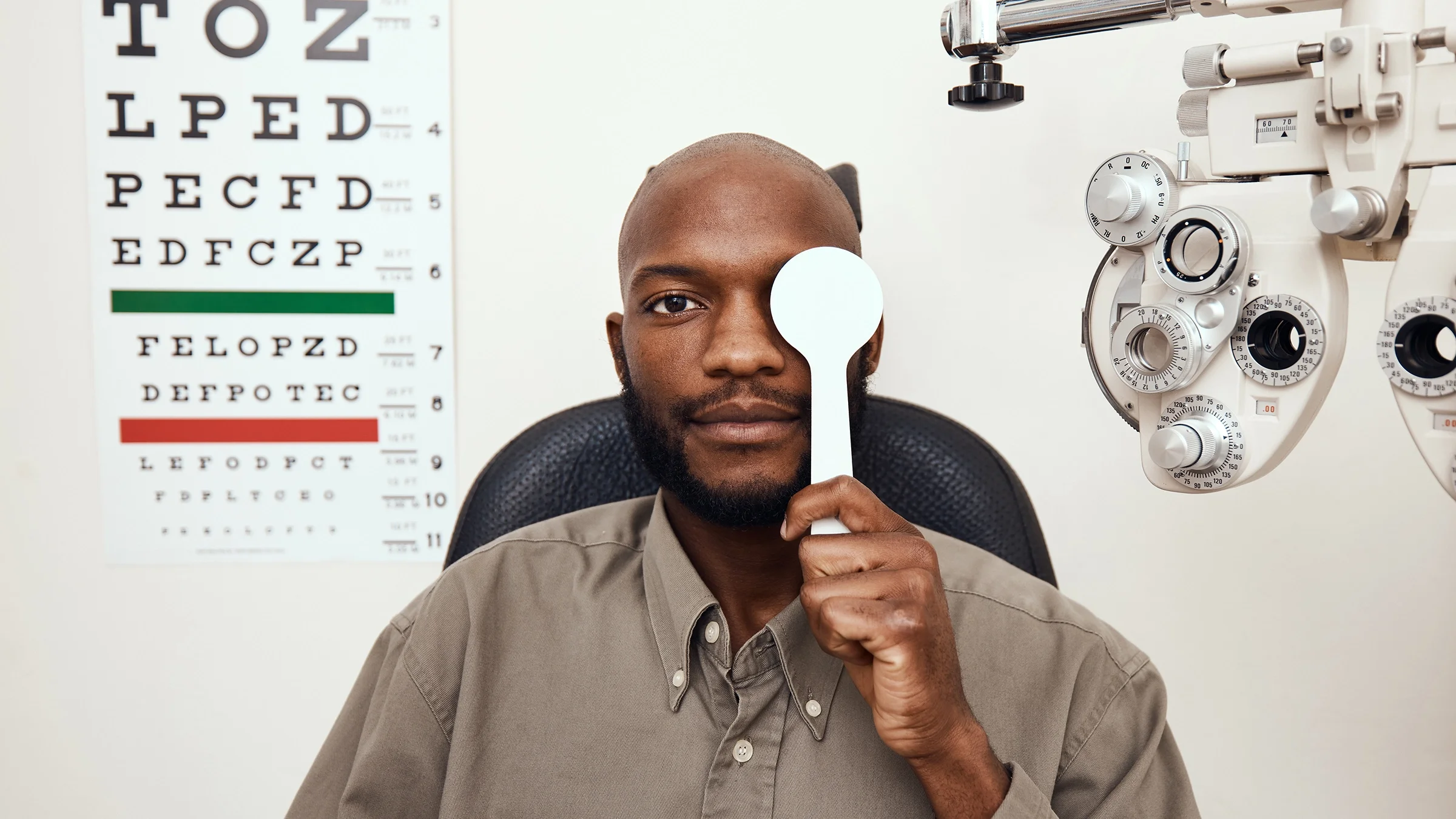 Man sitting in the eye exam room covering one eye. There is an eye chart behind him.