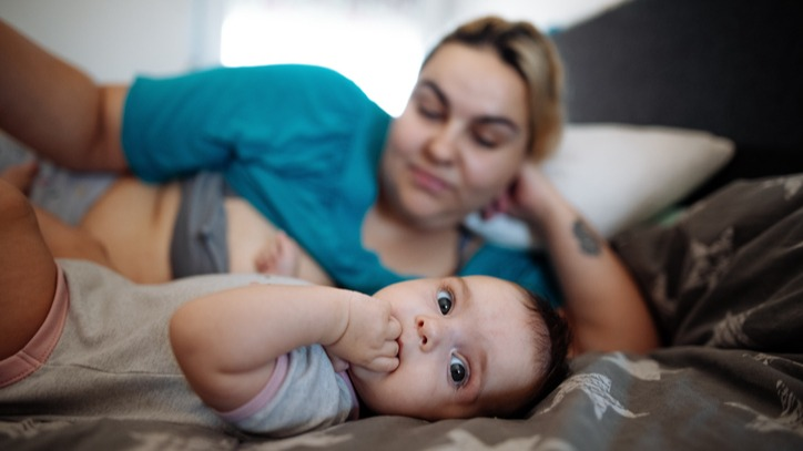A new mother looks at her baby while they both are lying on the bed. The baby is in focus looking at the camera with their fingers in their mouth and the mother is in the background out of focus.