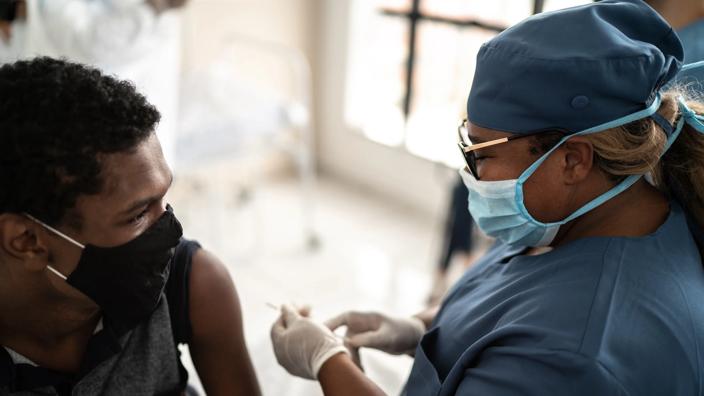 Teenager with a black face mask on getting the COVID-19 vaccine administered by a nurse in scrubs.