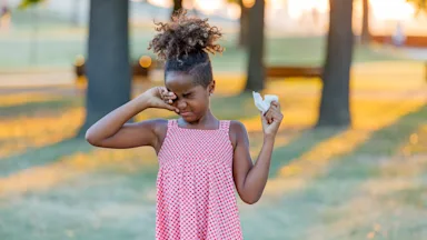 A child with allergies rubbing their eyes with one hand and holding a tissue in the other, at a park.
ProfessionalStudioImages/iStock via Getty Images 