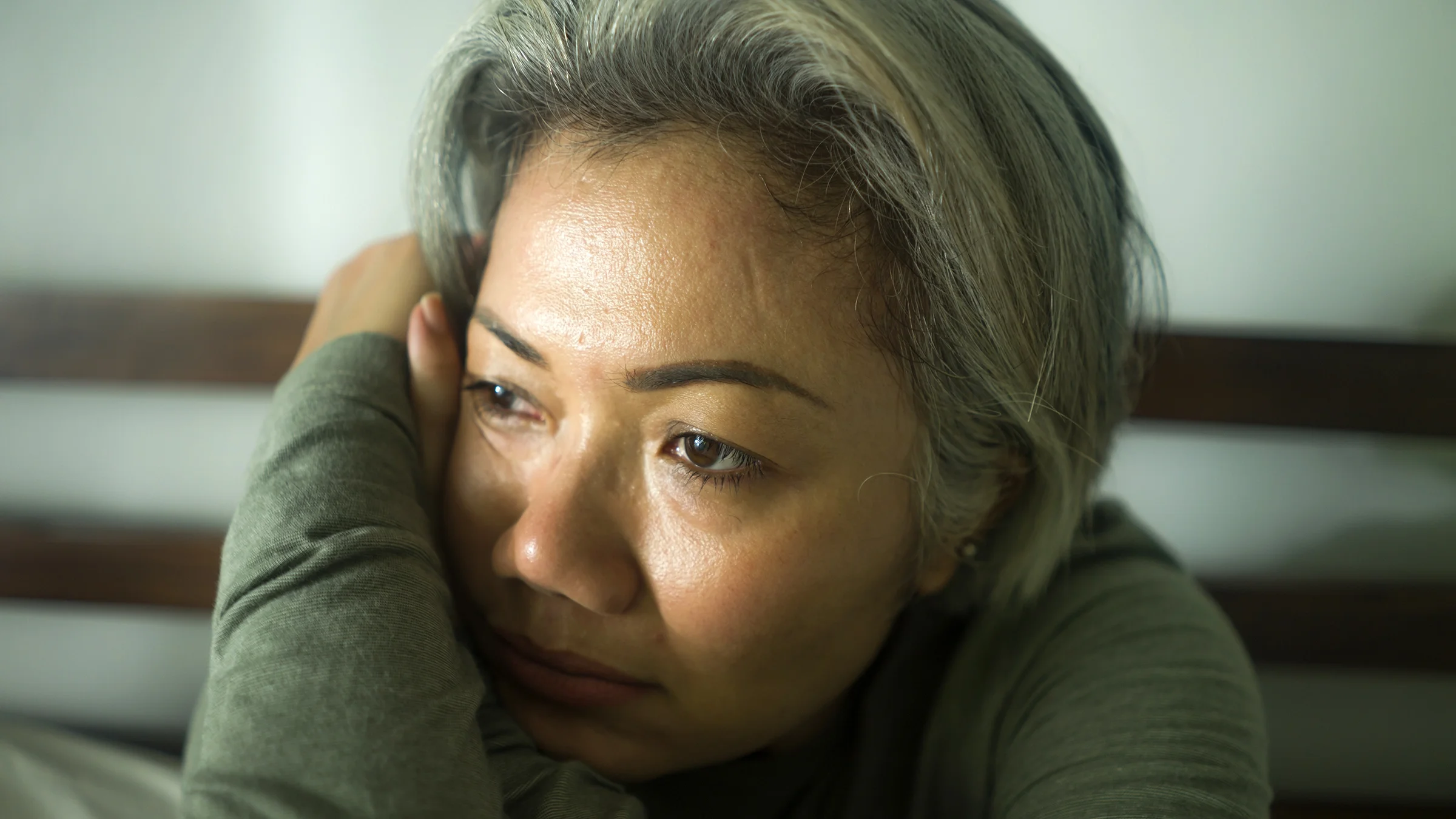Close-up of a mature woman resting her face on her arm while looking off into the distance with a sad expression.