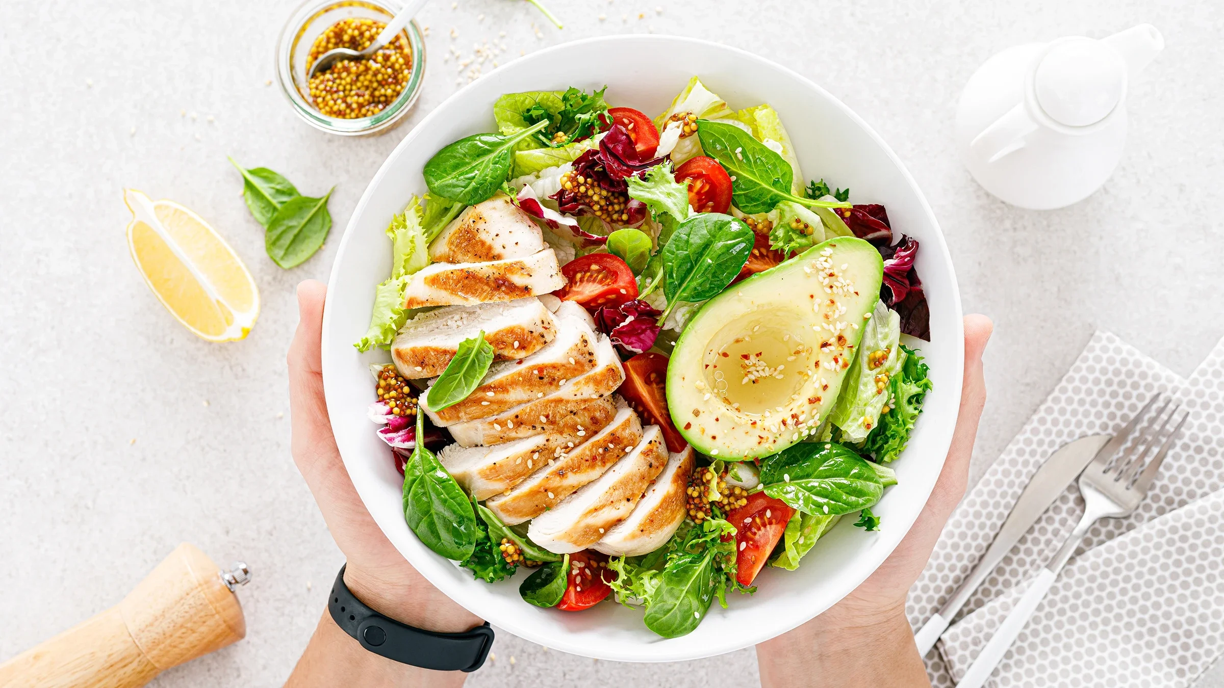 Overhead shot of a grilled chicken salad in a white serving bowl. A person's hands are holding it on either side of the bowl.