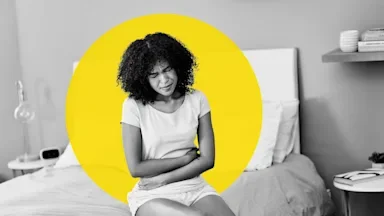 A black and white image of a woman sitting on the edge of her bed in pain. She is clutching her stomach. There is an added graphic yellow circle behind her.
LaylaBird/E+ via Getty Images
