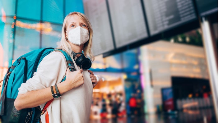 Woman with a backpack on walking around the airport with a face mask on. You can see the departure and arrival board behind her blurry in the background.
