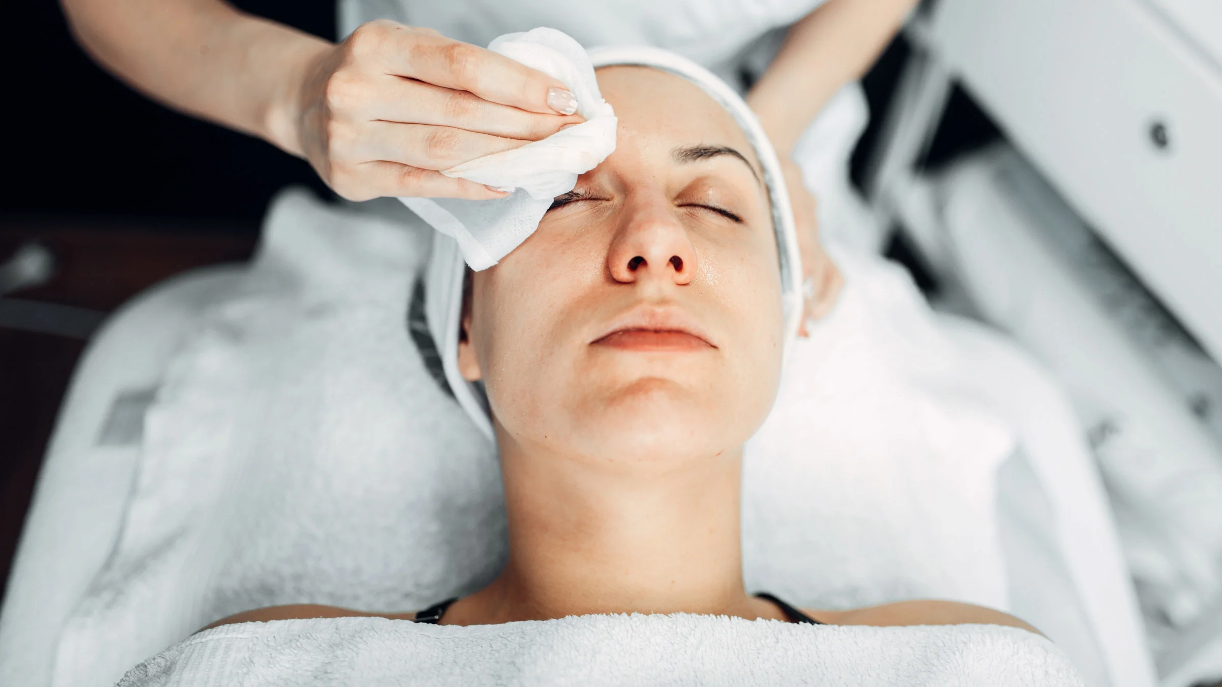 A young woman getting a facial with the esthetician's hands wiping her forehead with a white towel.