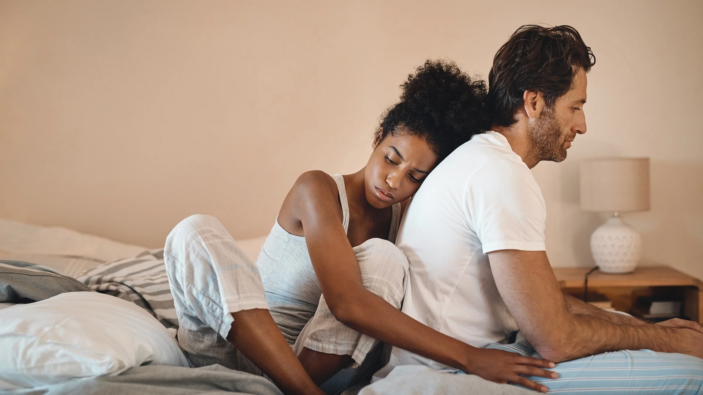 A woman hugging her partner as he sits on the edge of the bed. She is hugging him from behind. They are both sad and dealing with something heavy.